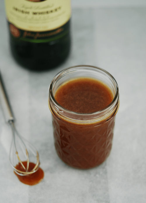 A jar of homemade Irish Whiskey Caramel Sauce with a bottle of Jameson Irish Whiskey behind it